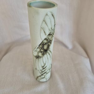 May include: A tall, cylindrical ceramic vase with a pale, off-white matte finish. The exterior features a dark, etched relief design of an insect with segmented wings and a textured, honeycomb-like body pattern extending vertically along the side.