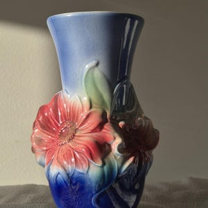 May include: A ceramic vase featuring a gradient of blue and white with a crackled glaze finish. The exterior is decorated with three-dimensional relief flowers in shades of pink, red, and green, extending from the base towards the flared rim.