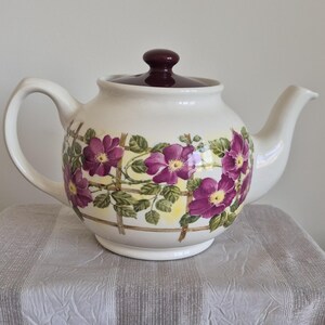 May include: A cream-coloured ceramic teapot featuring a dark maroon lid and a handle on the left side. The body is decorated with a wrap-around pattern of vibrant purple flowers and green leaves climbing along a brown wooden trellis design.
