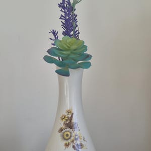 May include: A white ceramic vase with a narrow neck and a flared base, featuring a floral decal of yellow and brown sunflowers on the front. The vase contains a green succulent plant and a tall, thin sprig of purple artificial flowers.