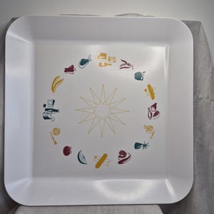 May include: A square white serving tray featuring a central gold starburst design surrounded by a circular pattern of colourful food illustrations including fruits, vegetables, cheese, bread, and wine bottles in shades of yellow, red, and teal.