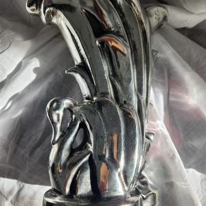 May include: A silver decorative vase featuring a swan design. The vase has a curved, flowing shape with a swan figure at the base. The vase is made of a shiny, reflective material, and the swan is depicted with its neck curved.