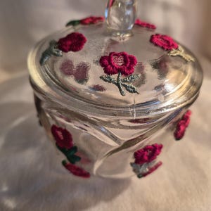 Peut inclure: Un pot en verre transparent avec un couvercle assorti, présentant un motif en relief tourbillonnant. La surface extérieure est ornée de petits motifs de roses rouges brodées avec des tiges et des feuilles vertes appliqués sur le verre.