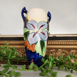 May include: A vintage ceramic vase with a floral design. The vase features a light blue flower with red accents, green leaves, and a blue base. It has two blue handles and is approximately 15 cm tall.