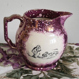 May include: A ceramic jug featuring a cream-coloured centre panel with a black line drawing of a person and two animals. The exterior is decorated with a purple and metallic gold sponge-painted pattern, including a handle and a pouring spout.