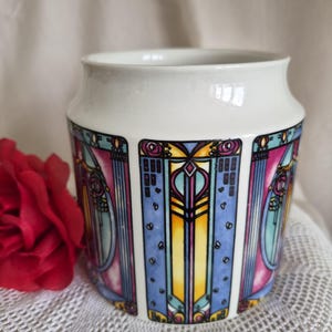 May include: A white ceramic jar featuring a repeating vertical pattern of stained glass-style panels in shades of blue, yellow, pink, and purple. The design includes geometric lines and circular motifs, with a smooth, wide opening at the top.