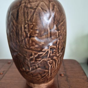 May include: A brown ceramic vase with a rounded, tapered shape and a smooth, glossy finish. The surface features a deep, embossed relief pattern depicting a landscape scene with trees and structures, rendered in varying shades of dark brown.