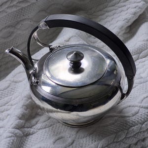 May include: A silver teapot with a black handle and a lid with a small knob. The teapot has a rounded body with horizontal ridges and a curved spout. The handle is arched and attached to the body of the teapot.