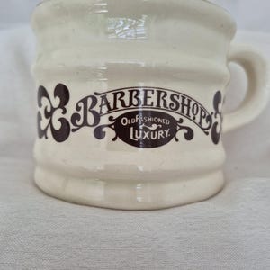 May include: A cream-coloured ceramic shaving mug with a ribbed exterior and a handle. The front features dark brown decorative scrollwork and the text "BARBERSHOP" arched above an oval containing the words "OLD FASHIONED LUXURY" in a serif font.