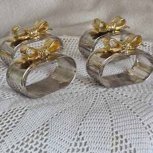 May include: Four oval-shaped napkin rings featuring a polished silver-tone finish and a decorative gold-tone bow accent attached to the top of each band. The set consists of two pairs of matching napkin rings with a smooth, reflective metallic surface.
