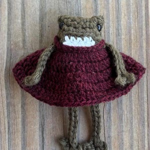 May include: A handmade crochet frog doll with a maroon dress and olive green limbs. The frog has a detailed head with black eyes and a white mouth. The doll is made of yarn and sits on a wooden surface.