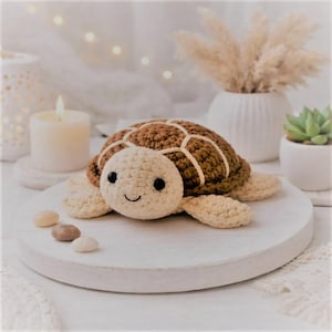 May include: A handmade crochet turtle with a brown shell and a cream-colored head and limbs. The turtle has black eyes and a smiling mouth. The turtle is sitting on a white wooden surface.