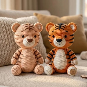 Puede incluir: Dos peluches de tigre hechos a mano con ganchillo. Uno es de color melocotón con rayas marrones, el otro es naranja con rayas negras. Ambos tienen vientre blanco, nariz marrón y ojos negros. Están sobre un sofá beige.