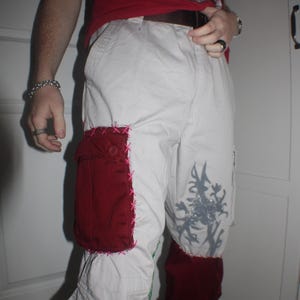 Puede incluir: Pantalones cargo blancos con parches burdeos y costuras rosas. Los pantalones presentan un diseño floral y un gráfico gris en la pierna. La persona lleva una camiseta roja y zapatillas blancas y burdeos.