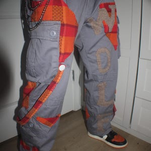 May include: Grey cargo trousers featuring orange and red plaid patches and the word "WOLF" in tan. They are accessorized with a silver chain and worn with black and orange trainers.