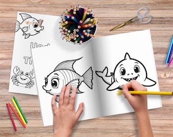 Può includere: Un libro da colorare aperto con disegni a tratto di pesci e uno squalo. Una mano sta colorando lo squalo con una matita gialla. Matite colorate, forbici e pastelli sono sparsi intorno al libro su una superficie di legno.