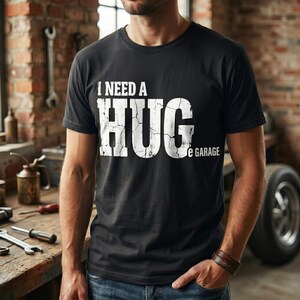 Op de afbeelding: Een zwart T-shirt met korte mouwen en de witte, verweerde tekst I NEED A HUG e GARAGE op de borst gedrukt. Het shirt heeft een ronde hals en een standaard pasvorm, met een grafisch ontwerp dat een gebarsten en versleten textuur vertoont.