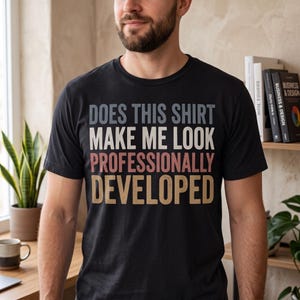 May include: A black short-sleeved t-shirt featuring the text "DOES THIS SHIRT MAKE ME LOOK PROFESSIONALLY DEVELOPED" printed in a stacked layout. The words are colored in shades of grey, white, light red, and tan with a distressed texture.