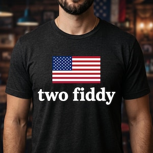 Peut inclure: Un t-shirt à manches courtes gris anthracite arborant un graphique du drapeau des États-Unis centré sur la poitrine. Sous le drapeau, le texte "two fiddy" est imprimé dans une police blanche, grasse et sans empattement. Col rond classique.