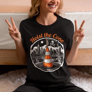 K&ouml;nnte beinhalten: Schwarzes T-Shirt mit einer Grafik, die einen Verkehrskegel mit einer Piratenflagge zeigt, auf der "Hoist the Cone" steht. Das Design beinhaltet eine Stadtlandschaft und den Text "Hoist the Cone" in Orange.
