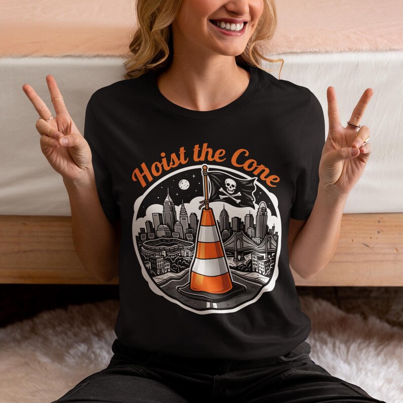 K&ouml;nnte beinhalten: Schwarzes T-Shirt mit einer Grafik, die einen Verkehrskegel mit einer Piratenflagge zeigt, auf der "Hoist the Cone" steht. Das Design beinhaltet eine Stadtlandschaft und den Text "Hoist the Cone" in Orange.