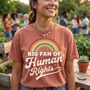 Peut inclure: Un t-shirt couleur terre cuite arborant un motif d'arc-en-ciel multicolore au-dessus du texte "BIG FAN OF Human Rights" dans une police rétro, agrémenté de petites étoiles et d'une ligne courbe soulignant le mot "Rights".