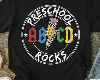 May include: A black t-shirt featuring a circular graphic with the text "PRESCHOOL ROCKS" in white. The center includes the letters "AB" in red and blue, "CD" in green and yellow, and a yellow pencil shaped like a lightning bolt with a checkered pattern.