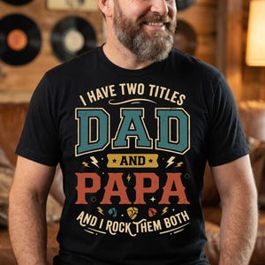 Peut inclure: Un t-shirt noir à manches courtes avec le texte I HAVE TWO TITLES DAD AND PAPA AND I ROCK THEM BOTH. Le motif présente les mots DAD en bleu sarcelle et PAPA en orange, agrémentés d'éclairs jaunes et de petites icônes de médiators.
