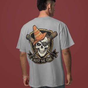 May include: A light gray t-shirt featuring a skull wearing an orange traffic cone and an eye patch. The skull is set against a shield with crossed baseball bats and the text "HOIST THE CONE".