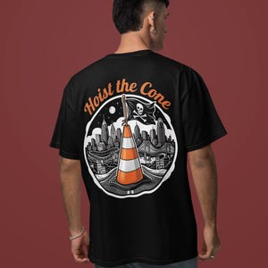K&ouml;nnte beinhalten: Schwarzes T-Shirt mit einem kreisf&ouml;rmigen Grafikdesign, das eine Stadtlandschaft, einen Verkehrskegel und eine Piratenflagge zeigt. Der Text "Hoist the Cone" ist in Orange &uuml;ber der Grafik angeordnet.