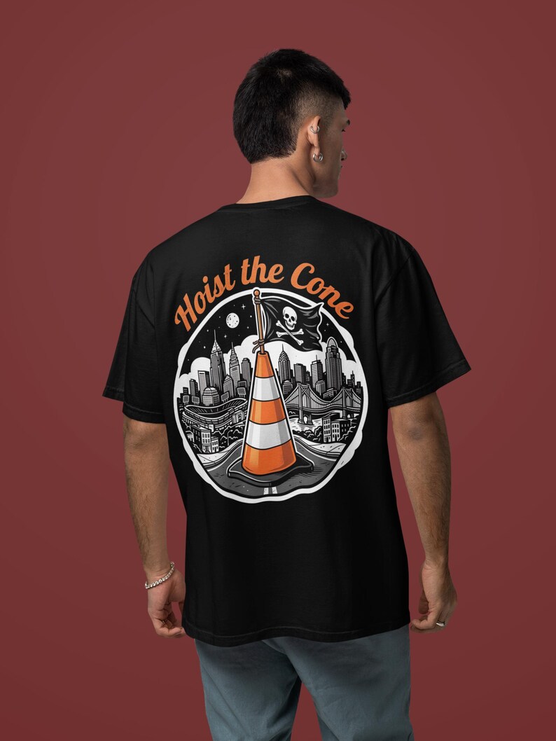 K&ouml;nnte beinhalten: Schwarzes T-Shirt mit einem kreisf&ouml;rmigen Grafikdesign, das eine Stadtlandschaft, einen Verkehrskegel und eine Piratenflagge zeigt. Der Text "Hoist the Cone" ist in Orange &uuml;ber der Grafik angeordnet.