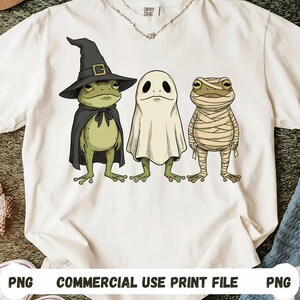 May include: White t-shirt featuring three cartoon characters: a frog in a witch hat and cape, a ghost, and a mummy. The frog is green, the ghost is white, and the mummy is wrapped in beige bandages. The text "COMMERCIAL USE PRINT FILE" is at the bottom.