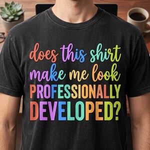 Peut inclure: Un t-shirt noir à manches courtes avec le texte multicolore does this shirt make me look PROFESSIONALLY DEVELOPED ? Le lettrage utilise un dégradé de couleurs orange, violet, bleu, jaune et vert sur quatre lignes de texte.