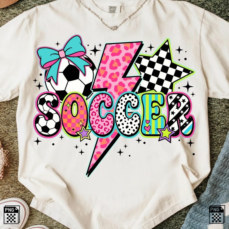 May include: White t-shirt with a colorful soccer-themed graphic. The design includes a soccer ball, a lightning bolt with a pink leopard print, stars, and the word "SOCCER" in various patterns and colors. A teal bow is above the soccer ball.