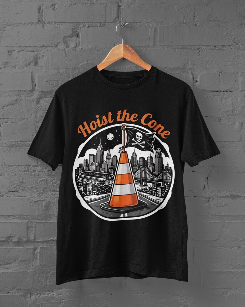K&ouml;nnte beinhalten: Schwarzes T-Shirt mit einer Grafik einer Verkehrskegel, einer Piratenflagge und einer Stadtlandschaft. Der Text "Hoist the Cone" ist &uuml;ber dem Bild angeordnet. Das Design verwendet Orange, Wei&szlig; und Schwarz.