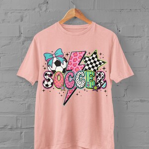 May include: A light pink t-shirt with a colorful graphic design. The design includes a soccer ball with a blue bow, a lightning bolt, stars, and the word "SOCCER" in various patterns and colors. The shirt is hanging on a wooden hanger.
