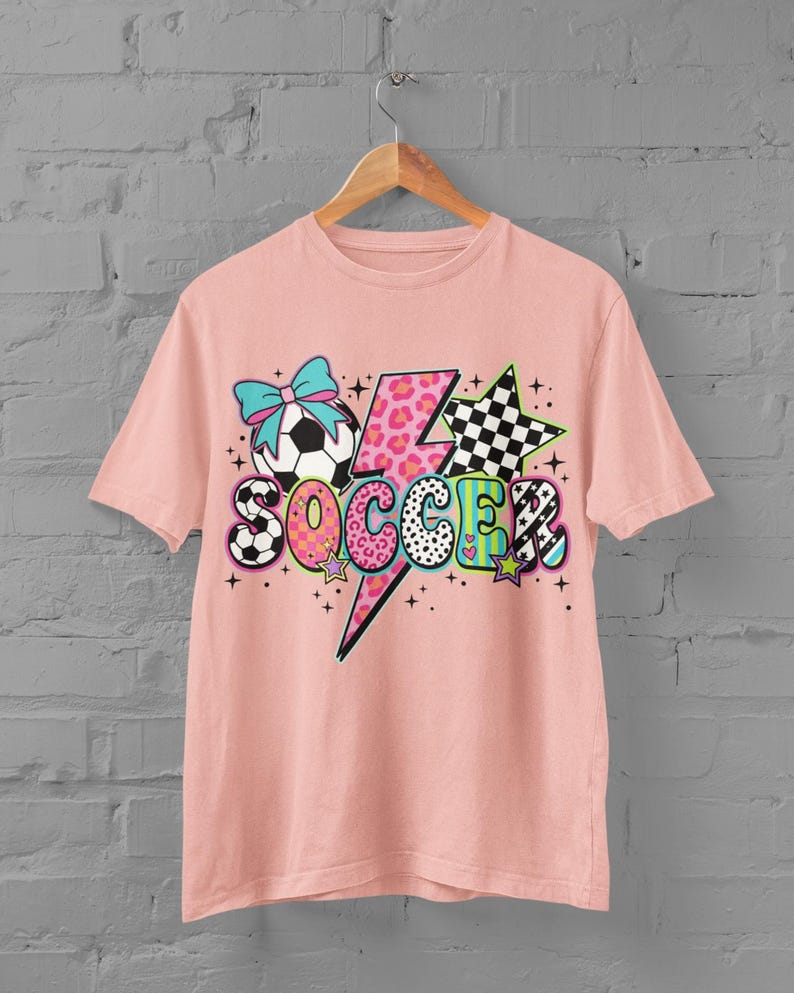 May include: A light pink t-shirt with a colorful graphic design. The design includes a soccer ball with a blue bow, a lightning bolt, stars, and the word "SOCCER" in various patterns and colors. The shirt is hanging on a wooden hanger.