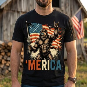 Peut inclure: T-shirt noir avec un motif de Bigfoot musclé portant des lunettes de soleil et un drapeau américain. Le Bigfoot fait un signe de la main rock and roll. Le mot "'MERICA" est imprimé en rouge, blanc et bleu. Design patriotique.