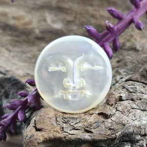 Op de afbeelding: Een ronde, iriserende witte parelmoeren cabochon met een uitgesneden, gestileerd menselijk gezicht met gesloten ogen, een gedefinieerde neus en een kleine mond. Het oppervlak heeft een gladde, gepolijste afwerking met subtiele parelmoerkleuren.