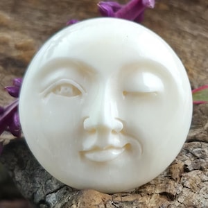 May include: A circular, cream-coloured moon face with a winking eye and smiling expression. The moon face is carved with detailed features and rests on wood with purple flowers.