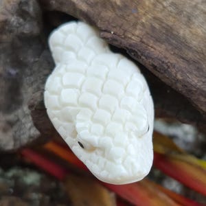 May include: A carved white snake head figurine featuring detailed scale patterns across the surface and small, dark, circular eyes. The sculpture is shaped with a rounded snout and a textured top, showcasing a smooth, off-white finish throughout.