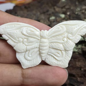 Op de afbeelding: Ivoor kleurige vlindersnijwerk met gedetailleerde vleugelpatronen en lichaamssegmenten. De vlinder wordt in een hand gehouden, wat het ingewikkelde reliëfwerk laat zien. Kan een broche of hanger zijn.