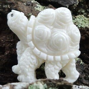 May include: A white carved turtle figurine featuring a shell decorated with multiple raised rose patterns. The turtle has a smooth, polished surface with detailed textures on its legs and head, showcasing a monochromatic design throughout the piece.
