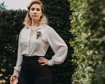 Puede incluir: Una mujer con una blusa blanca abotonada con un alfiler floral, pantalones negros de cintura alta y un collar. Está de pie frente a un seto verde.