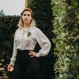 Op de afbeelding: Een vrouw draagt een wit overhemd met knopen en een bloemspeld, een zwarte broek met hoge taille en een ketting. Ze staat voor een groene haag.