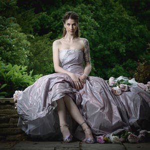 K&ouml;nnte beinhalten: Eine Frau in einem flie&szlig;enden, helllila Kleid sitzt auf einer Steinmauer, umgeben von rosa Blumen. Sie tr&auml;gt eine zarte silberne Halskette und hat ein Tattoo auf ihrem Arm.