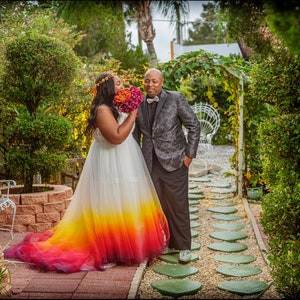 African American bride and groom stand in garden. The bride wears a wedding dress dipdyed bright colours of yellow, red and purple