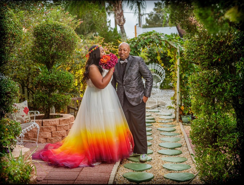 African American bride and groom stand in garden. The bride wears a wedding dress dipdyed bright colours of yellow, red and purple