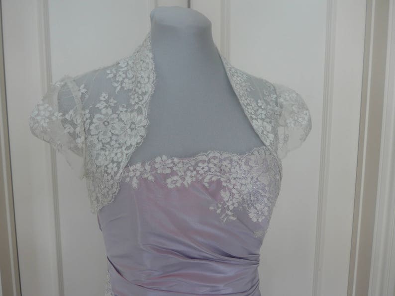 K&ouml;nnte beinhalten: Ein lavendelfarbenes Satin-Kleid mit einer wei&szlig;en Spitzen&uuml;berlage. Die Spitzen&uuml;berlage ist eine Bolero-Jacke mit kurzen &Auml;rmeln.