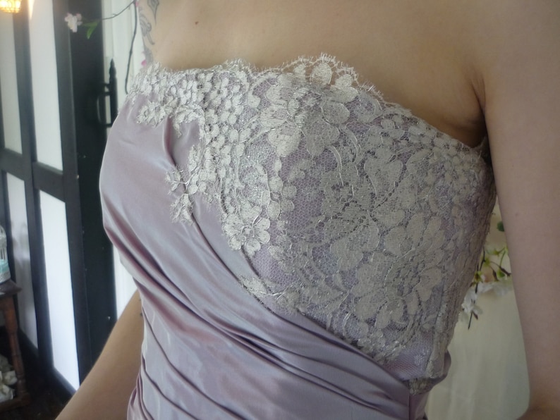 K&ouml;nnte beinhalten: Ein tr&auml;gerloses Kleid mit einem Lavendel-Satin-Oberteil und einer wei&szlig;en Spitzen-&Uuml;berlagerung. Das Oberteil ist gerafft und die Spitze ist zart und filigran.